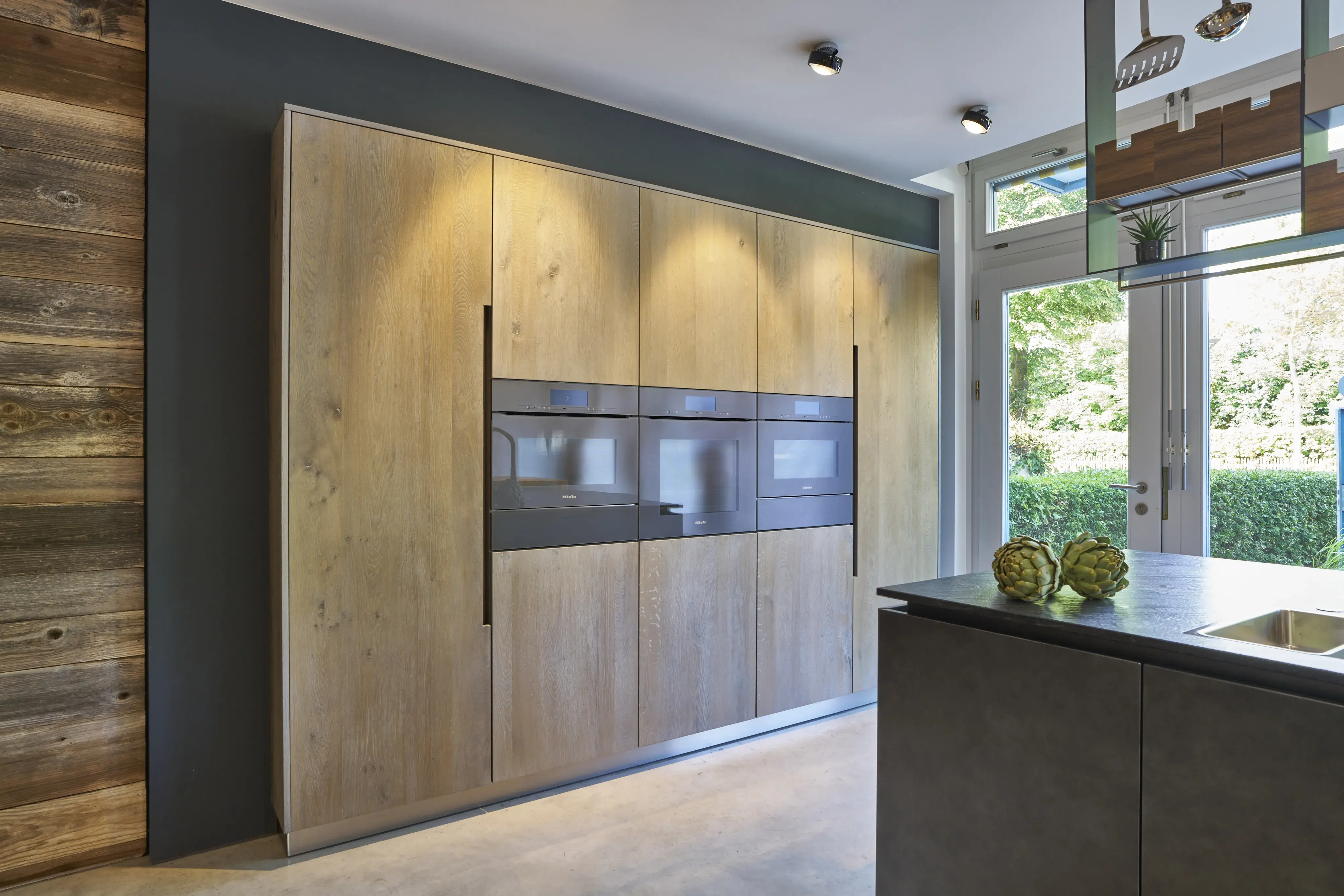 Moderne Küche mit Holzfronten, schwarzer Arbeitsplatte und großen Fenstern im Hintergrund.