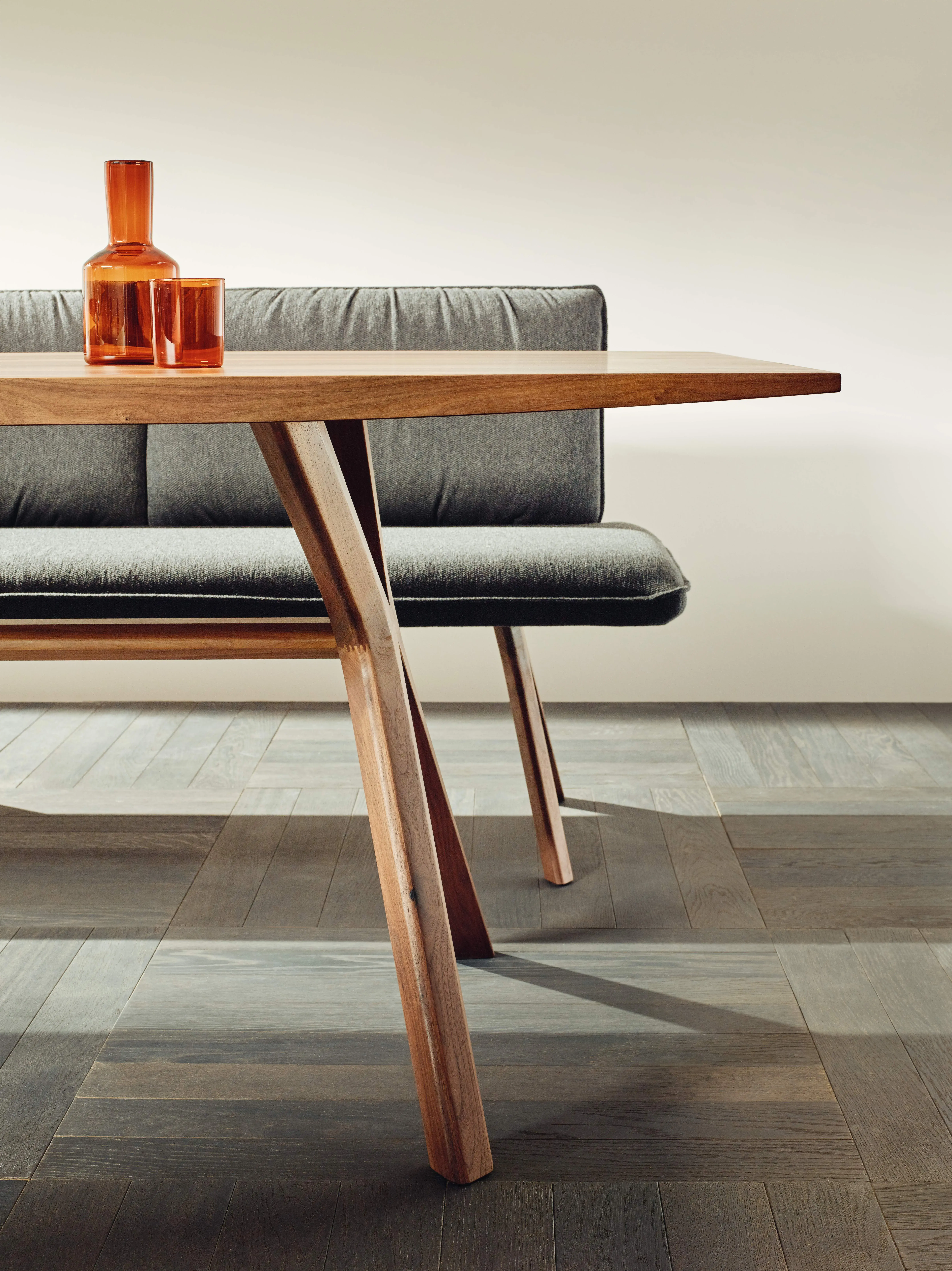Holztisch mit orangen Glasvasen, dahinter eine graue Polsterbank auf Holzfußboden.