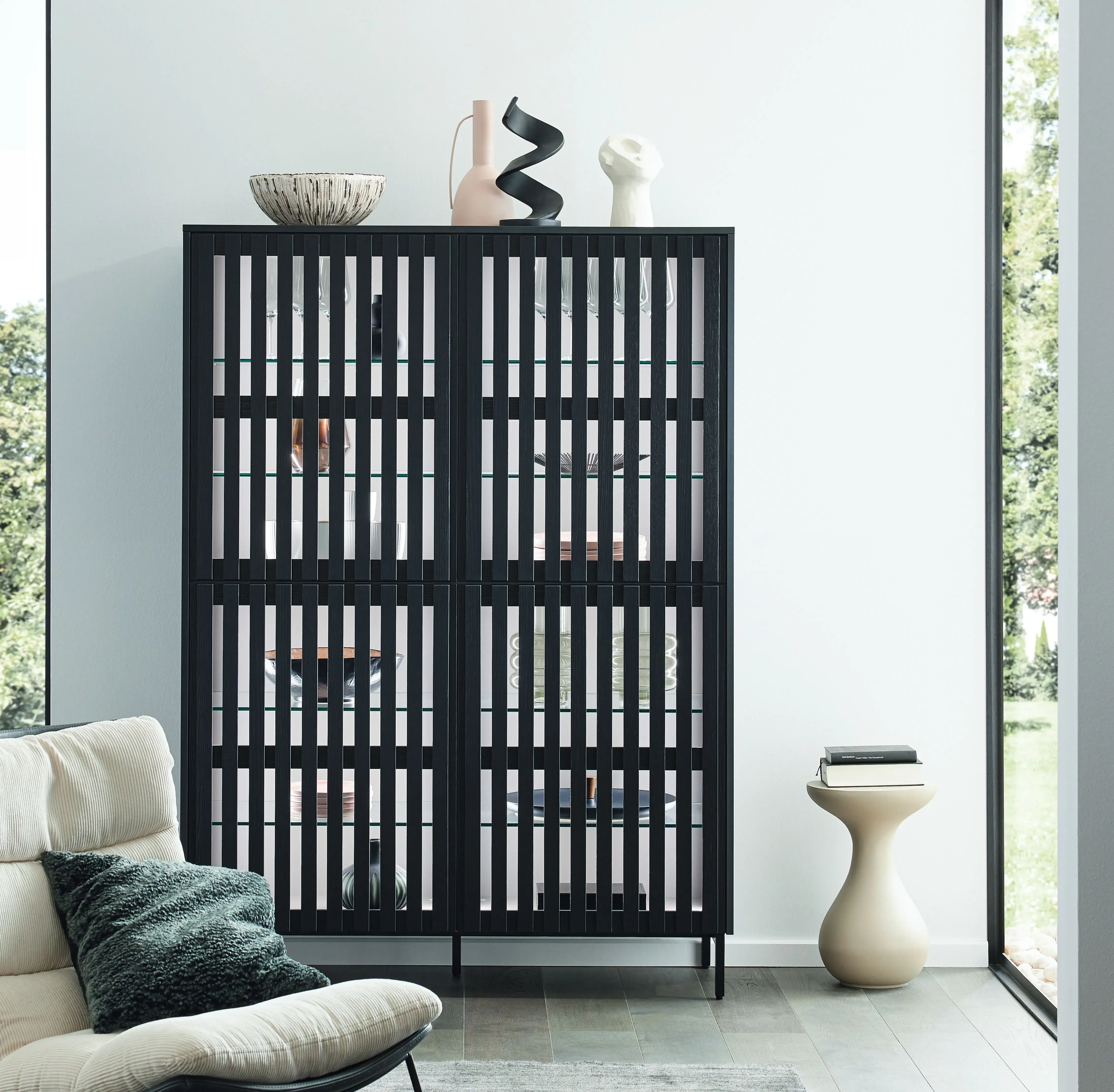 Schwarzer Schrank mit Zierobjekten davor, helle Wand und großes Fenster.